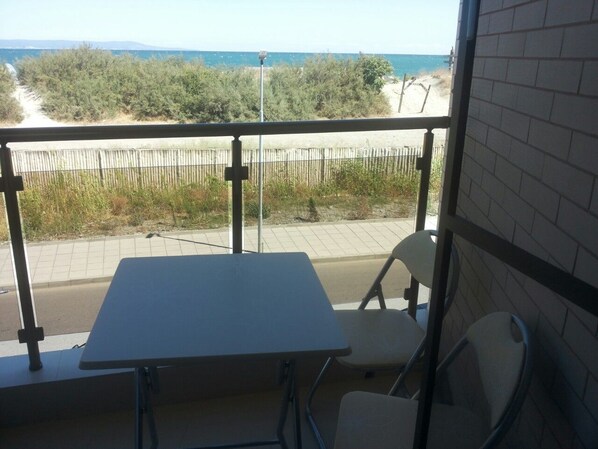 Un balcón con una mesa y dos sillas, con vista a una playa con cercado y vegetación.