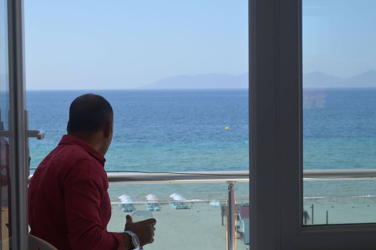 A man seated by a window overlooking a beach with umbrellas and lounge chairs.