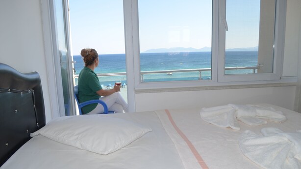 A hotel room with a bed, a window with a view of the sea, and a person sitting by the window. - Yasmin Hotel (Ayvalik)
