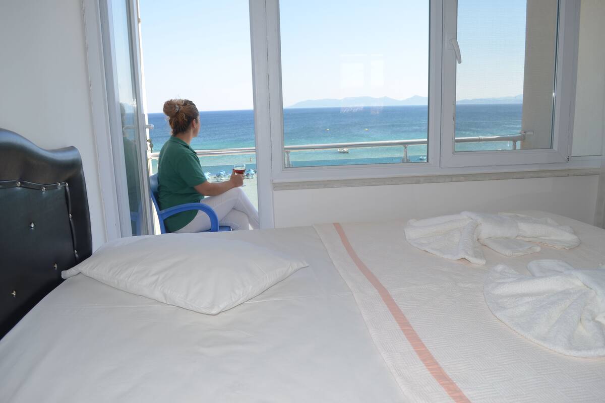 A hotel room with a bed, a window with a view of the sea, and a person sitting by the window.