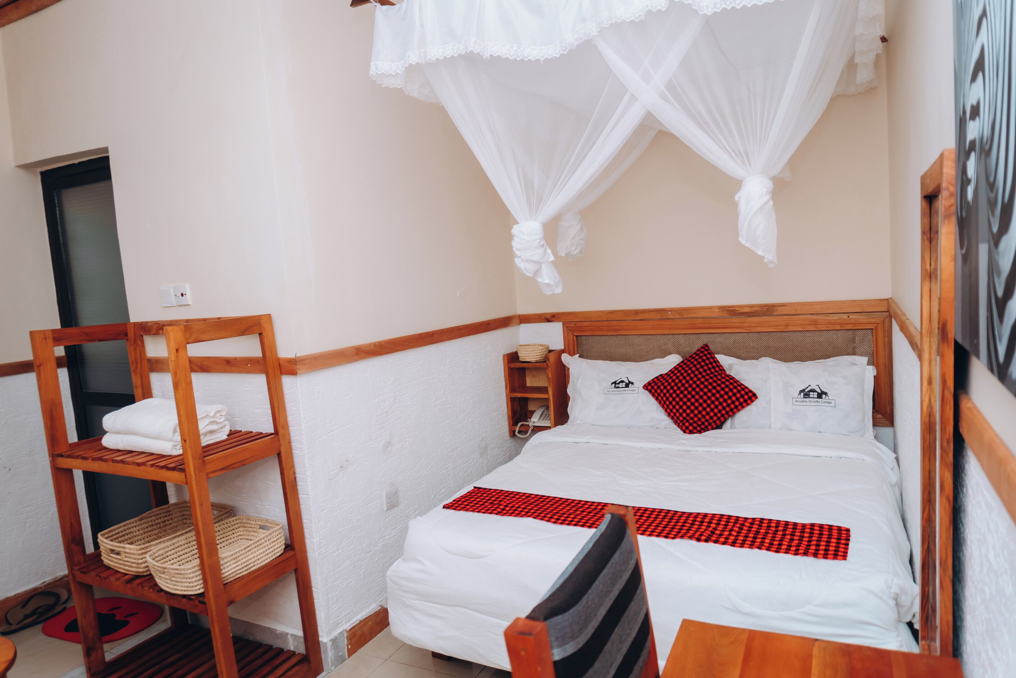 A bedroom with a wooden headboard, a bed with white bedding and red accents, a wooden shelf with baskets, and a mosquito net above the bed.