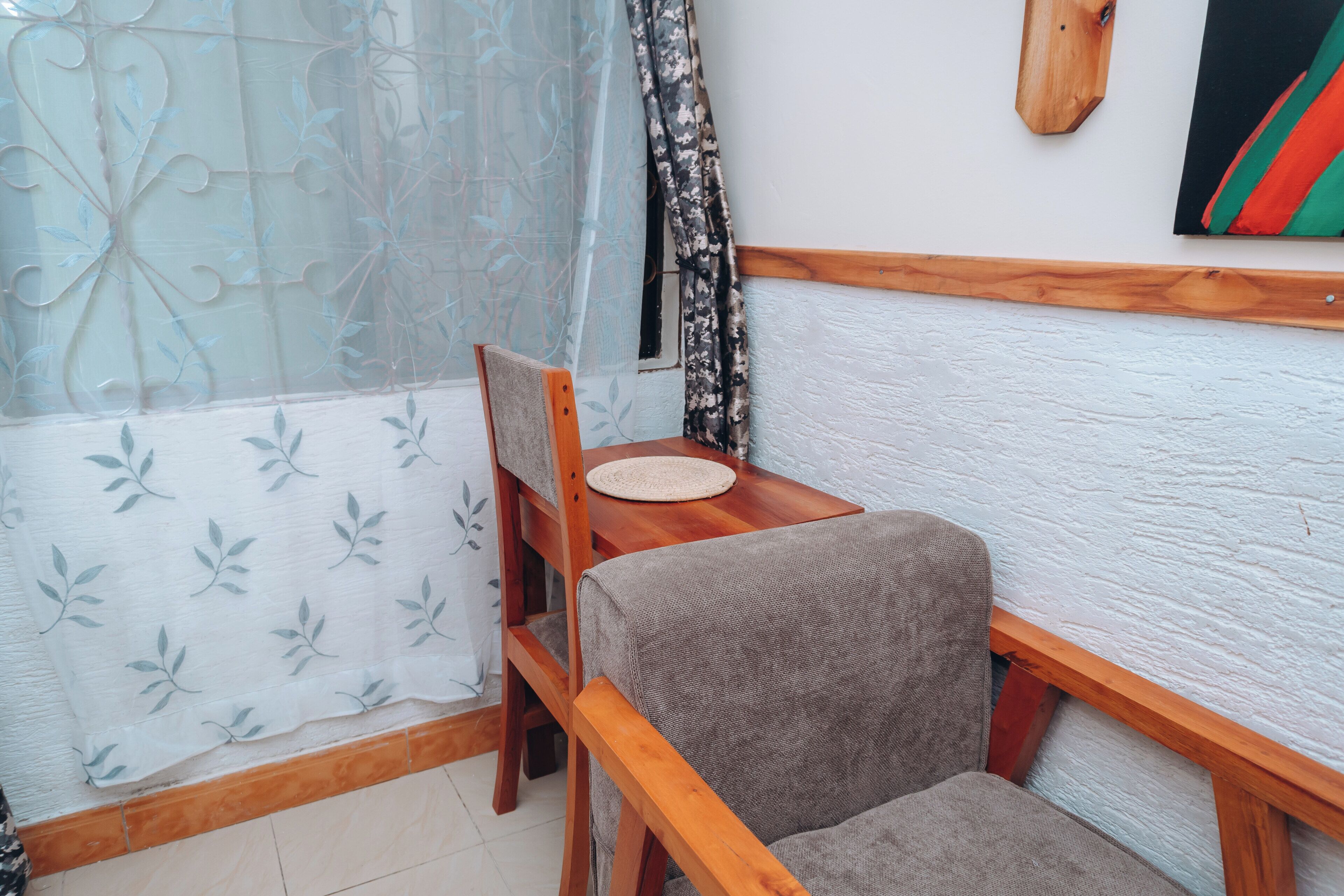 A room with a wooden chair, a small table, a window with a sheer curtain, and a wall-mounted painting.