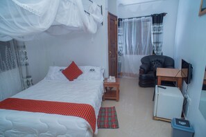 A bedroom with a bed, a chair, a small table, a refrigerator, and a window with curtains. - Arusha Giraffe Lodge (Arusha)