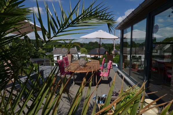Terraza en la azotea con una mesa y sillas de madera, rodeada de macetas con plantas y sombrillas.