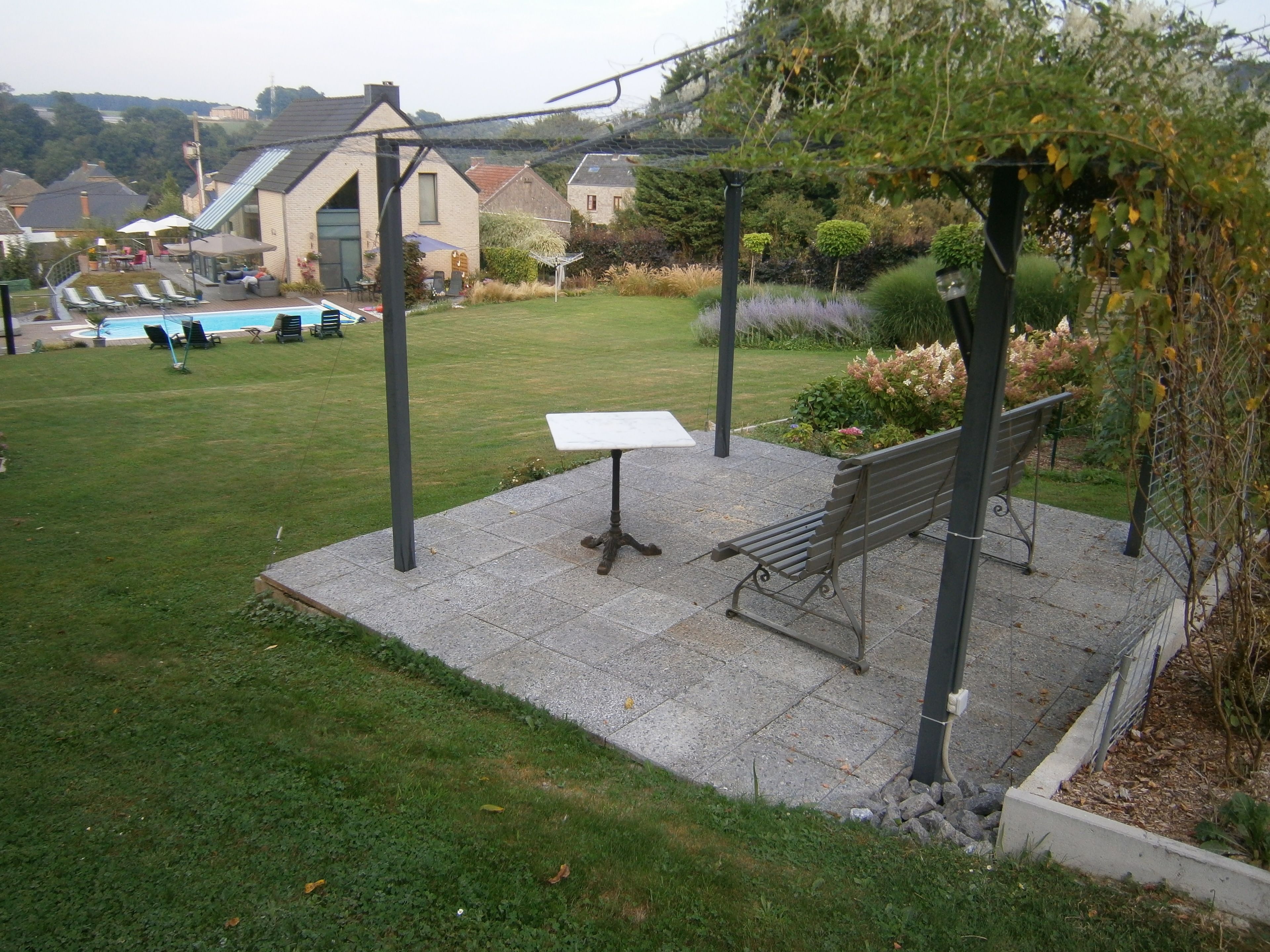 Une terrasse avec un banc et une table, entourée d’un jardin où poussent différentes plantes, et une piscine en arrière-plan.