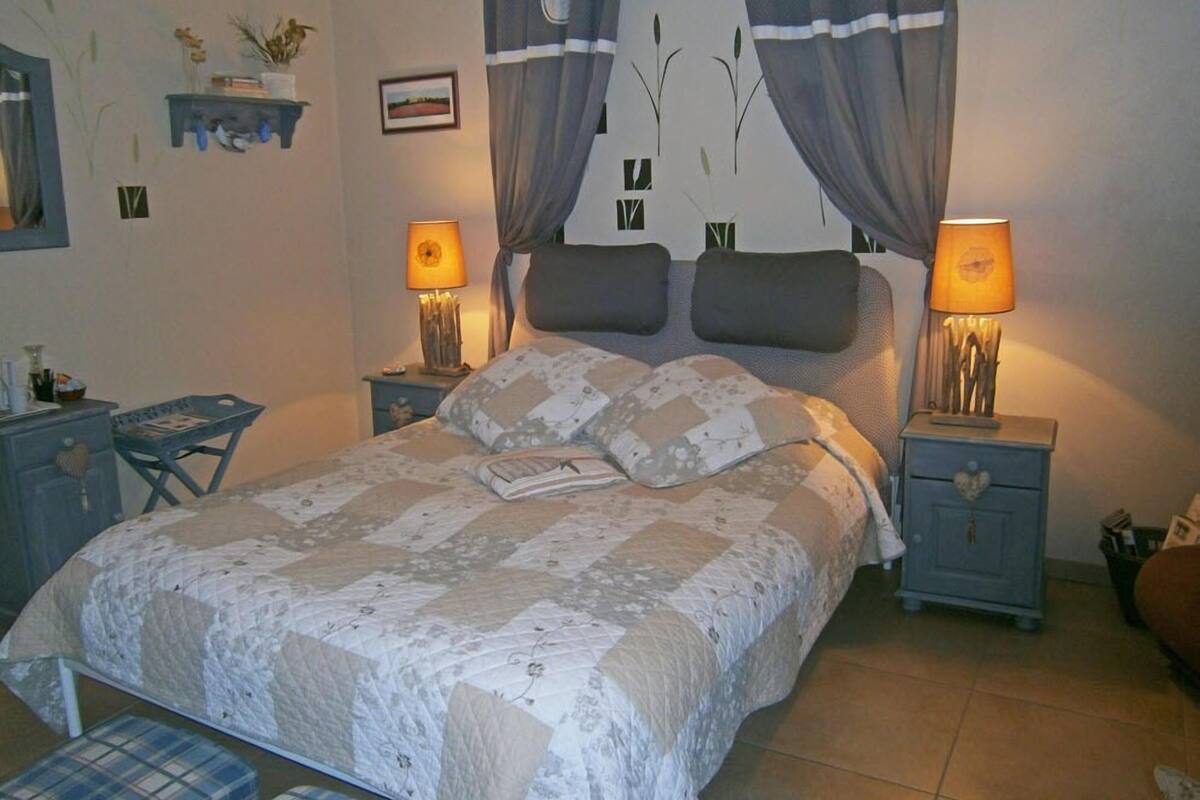A bedroom with a bed, two bedside tables, a nightstand, a chair, and a wall with a picture and a shelf with decorative items.
