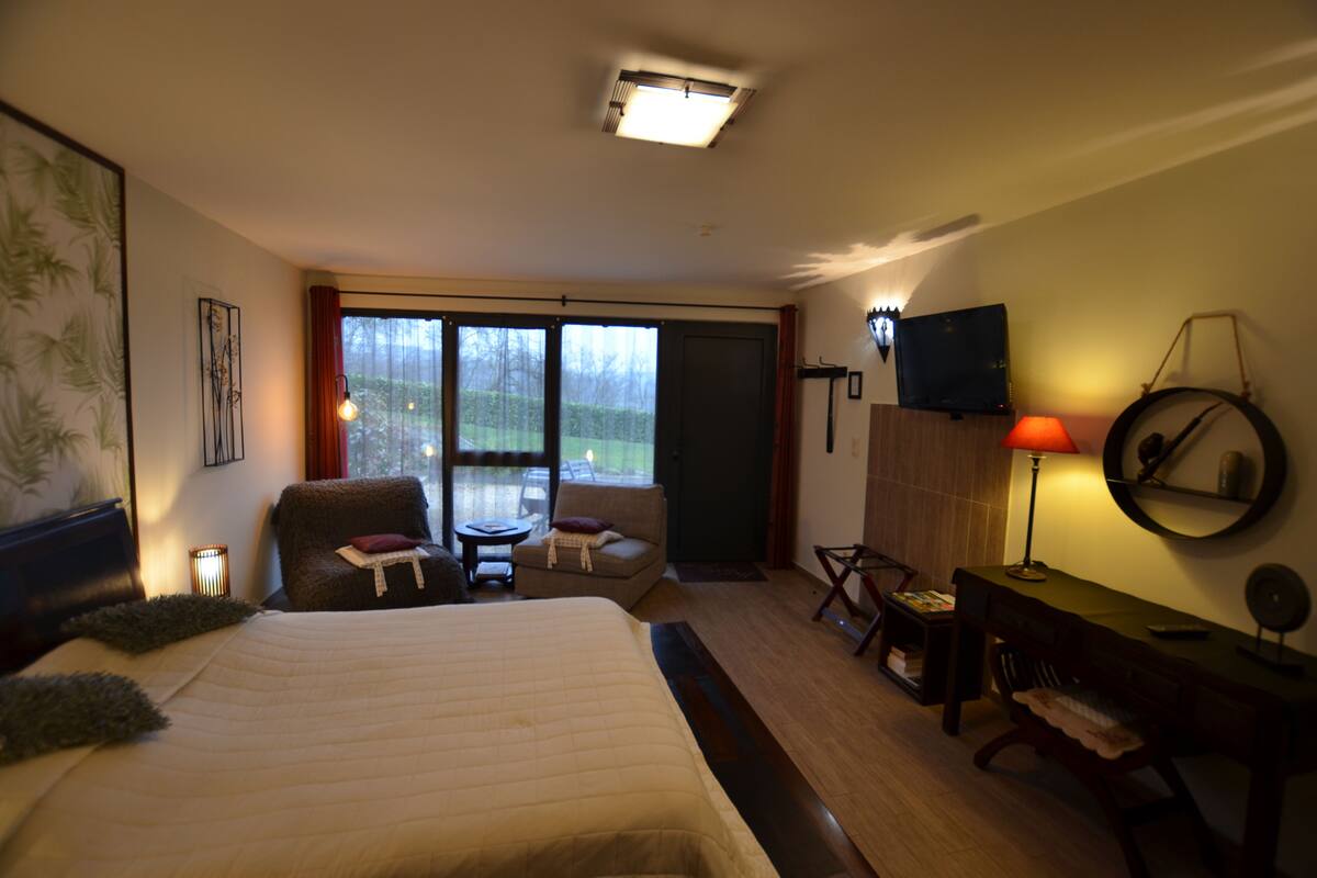 A hotel room with a large bed, a desk with a chair, a small table, a TV, and a view of the outdoors through the window.