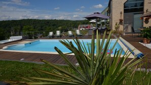 Una piscina con sillas de descanso y sombrillas, rodeada por una terraza de madera y vegetación.