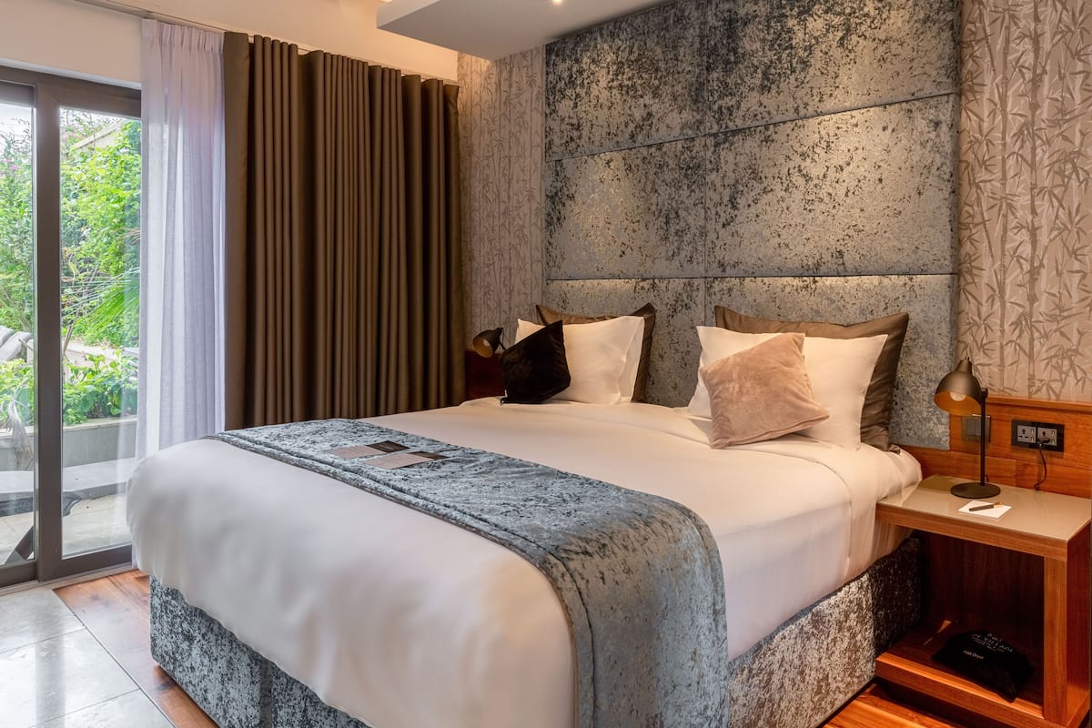 Quarto de hotel moderno com uma cama grande, mesa de cabeceira, abajur e vista para o verde pela janela.
