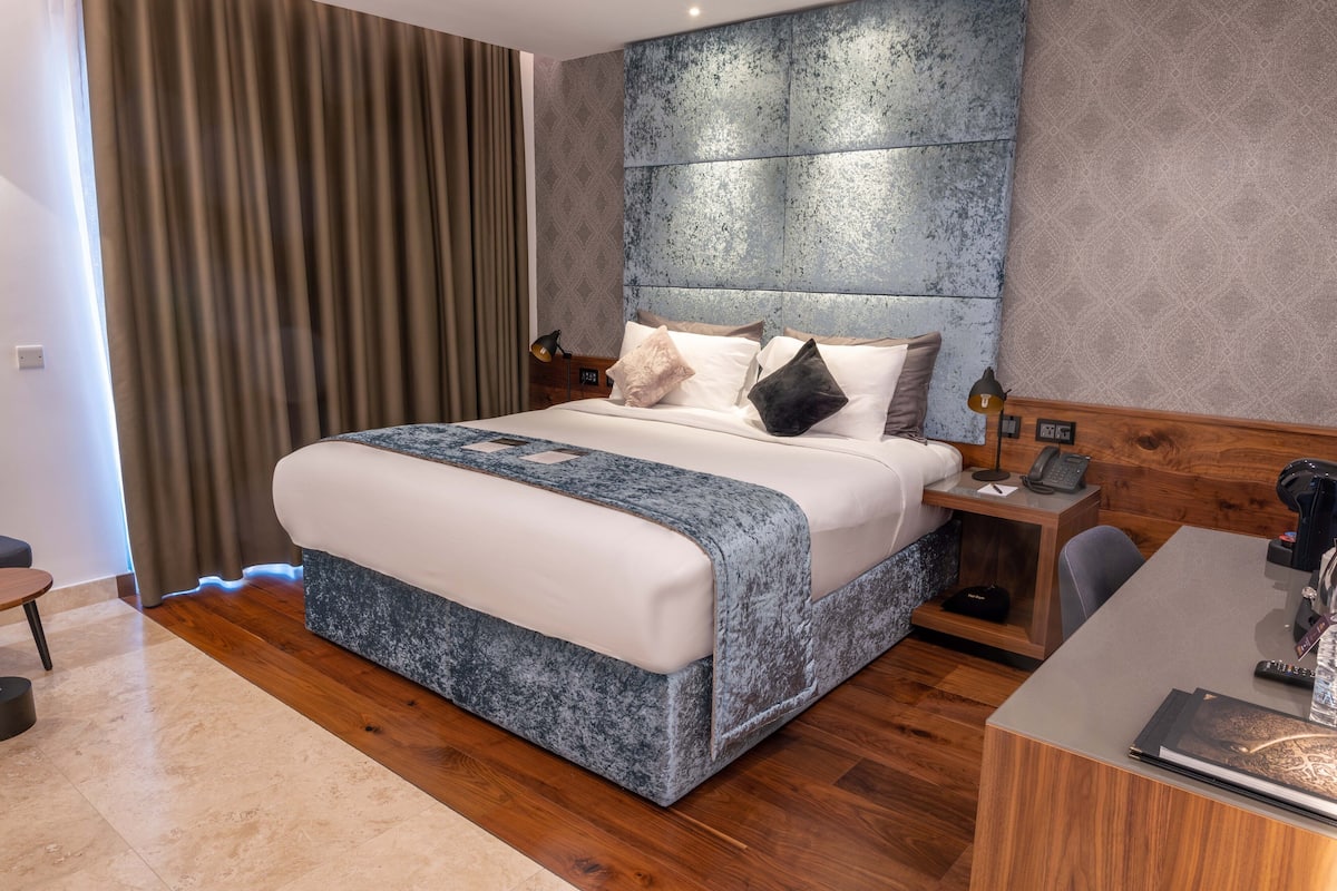 Quarto de hotel moderno com uma cama grande, piso de madeira, escrivaninha com cadeira e mesa de cabeceira com abajur e telefone.