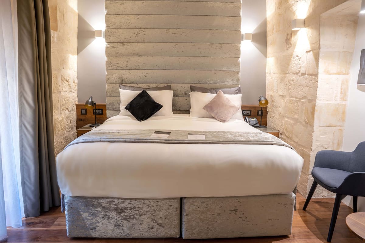 Uma cama grande com cabeceira de pedra, duas mesas de cabeceira com abajures e uma cadeira.