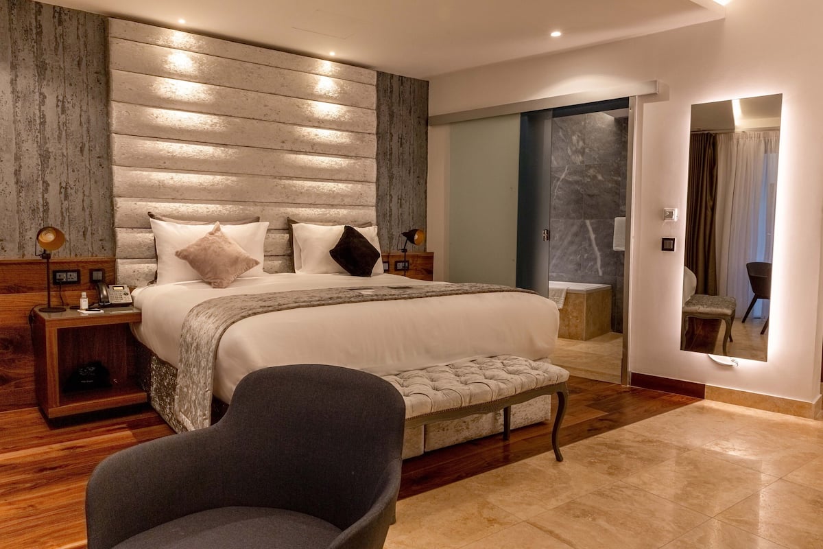 Quarto moderno com cama grande, cabeceira de madeira, luminárias de cabeceira, um banquinho e um espelho.