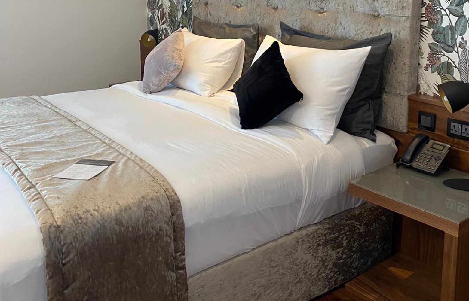 A neatly made bed with a white comforter and pillows, a wooden nightstand with a telephone, and a wall with a floral pattern.