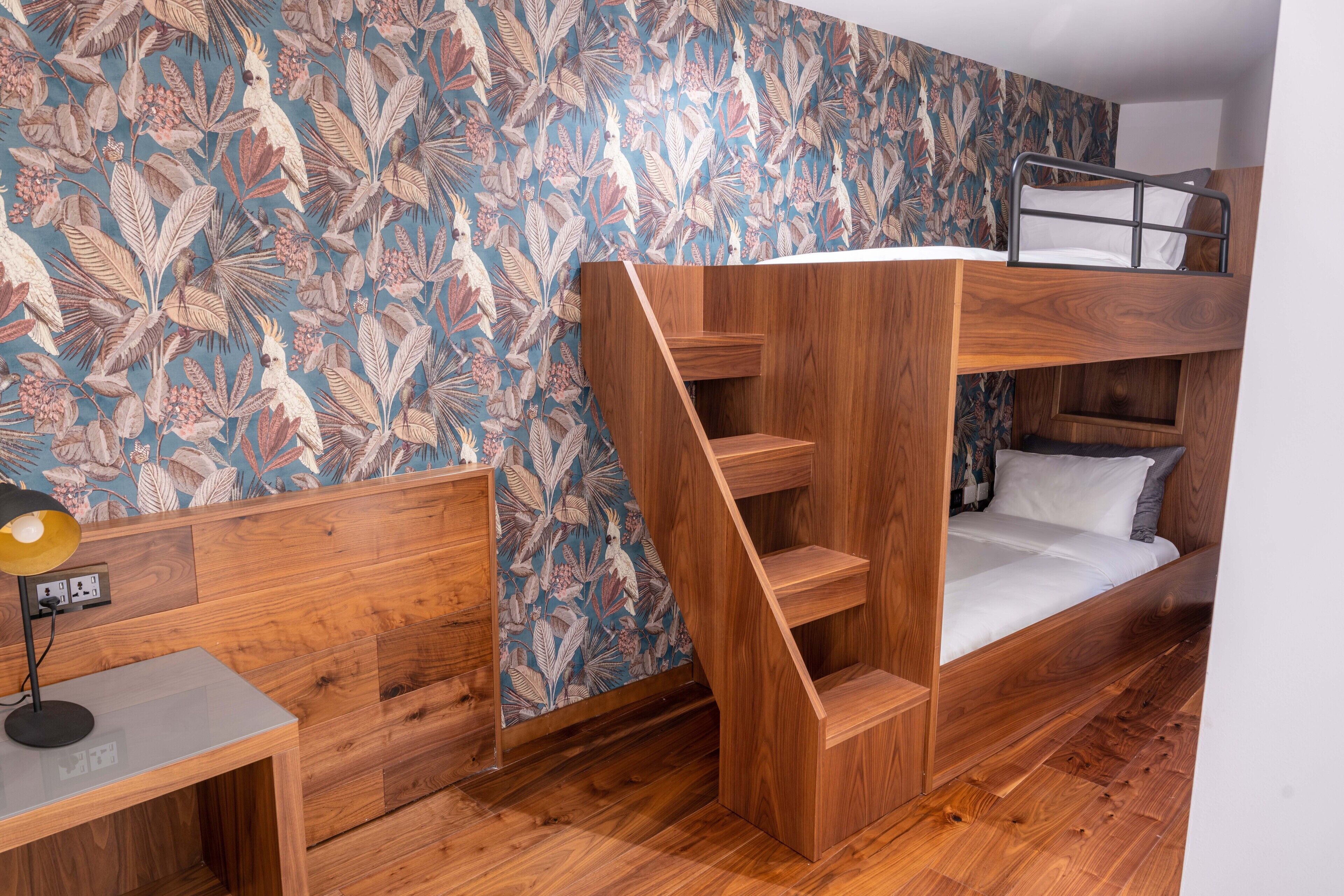 A bunk bed with a wooden ladder, a desk with a lamp, and a wall with a floral pattern.