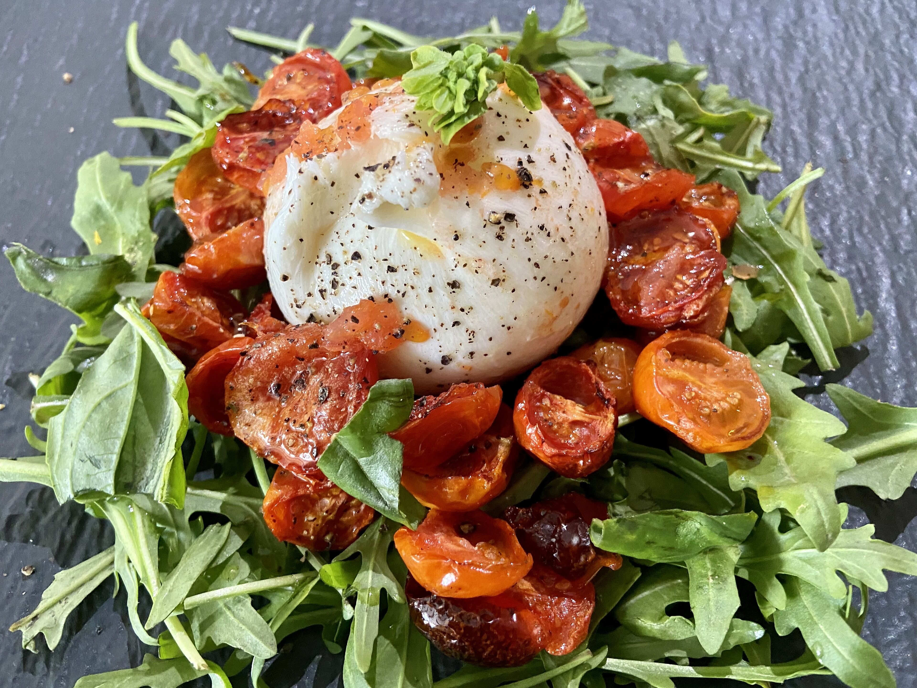 Un plato con un huevo escalfado, tomates asados y rúcula sobre una pizarra.