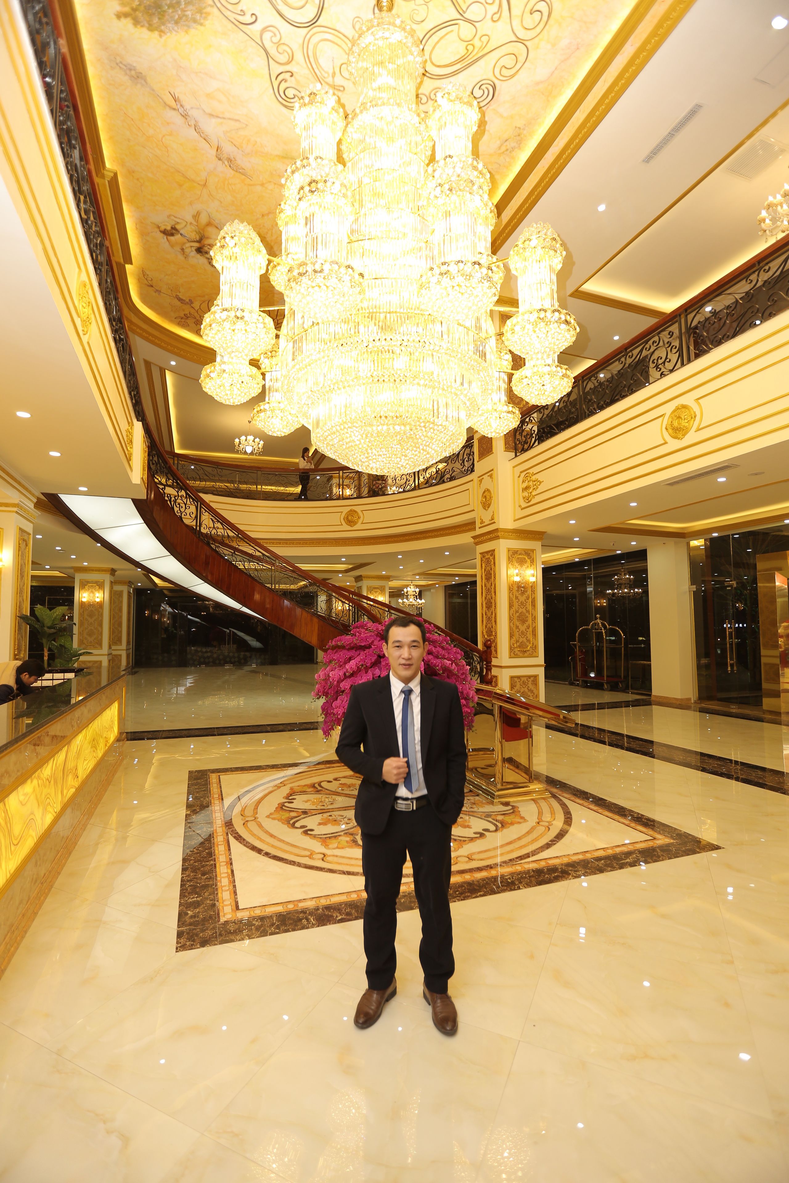 Ein Mann in einem Anzug steht in einer Empfangshalle mit einem großen Chandelier, Marmorböden und einer prachtvollen Treppe.