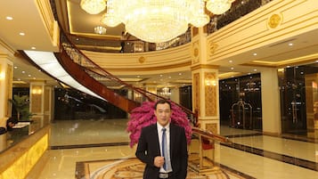 A man in a suit standing in a lobby with a large chandelier, marble floors, and a grand staircase.