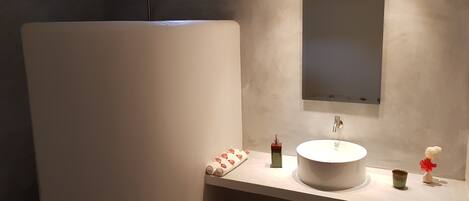 A modern bathroom with a white sink, a mirror, and a shower. There is a television mounted on the wall and a small shelf with toiletries.