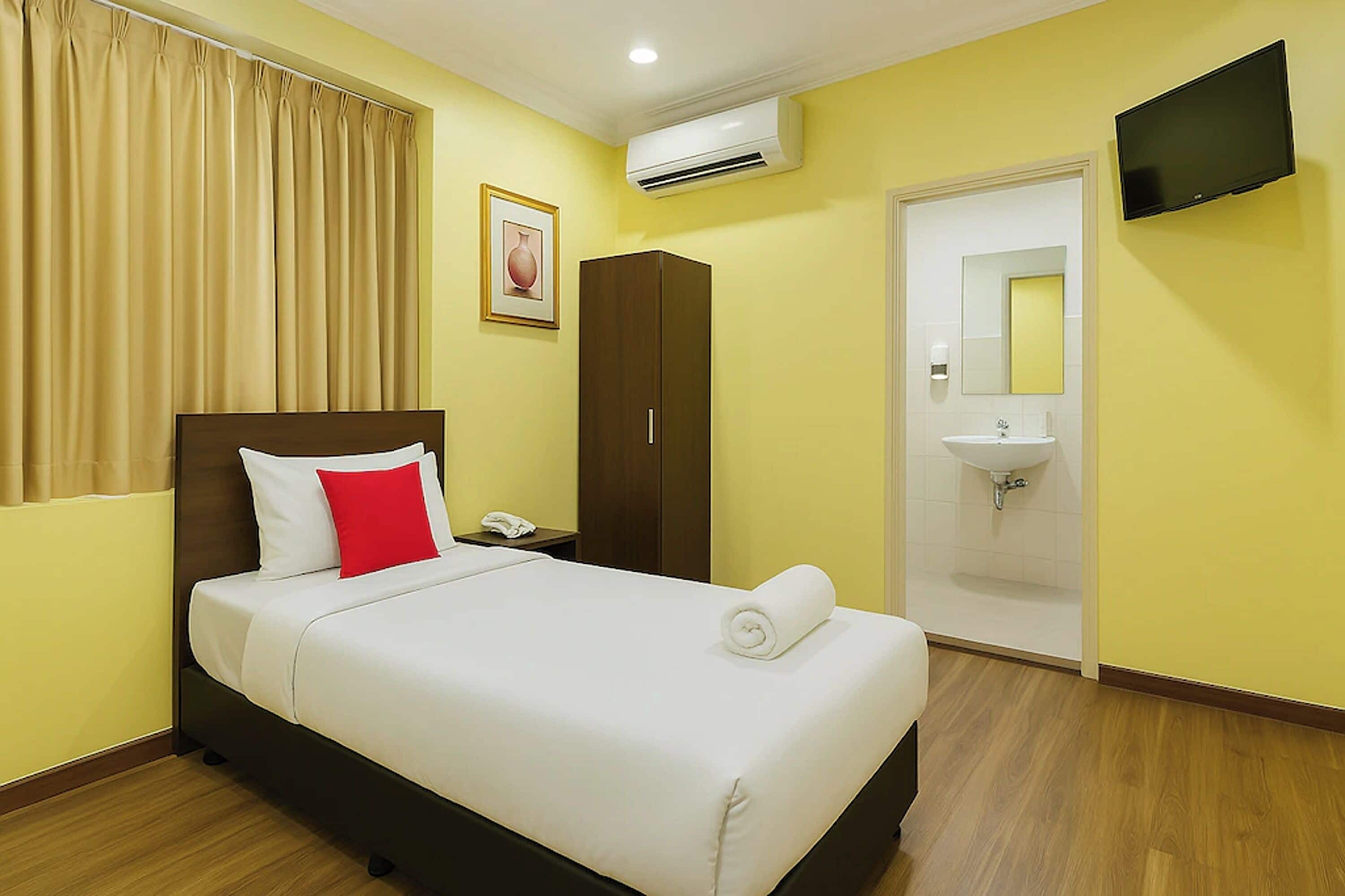 A hotel room with a bed, a wardrobe, a bedside table, a telephone, an air conditioning unit, a television, and a bathroom with a sink and mirror.