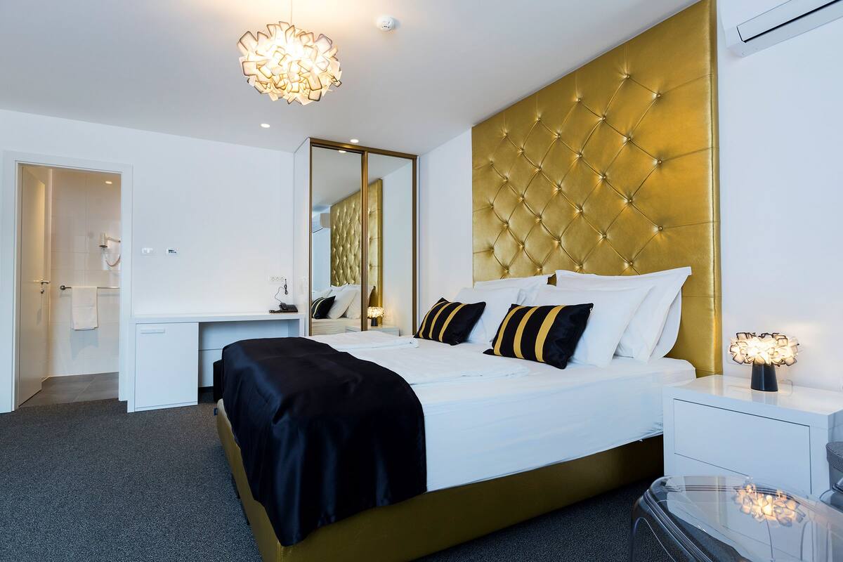 A modern hotel room with a large bed, a gold headboard, a bedside table with a lamp, and a large mirror.