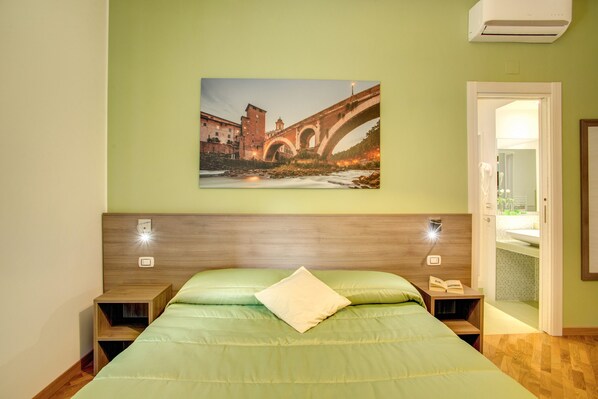 Una habitación de hotel con una cama, una mesita de noche, una lámpara de lectura y un cuadro de un puente.