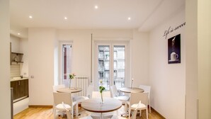 Una cocina moderna con armarios blancos y encimera de madera. Hay dos mesas de comedor blancas con sillas, y un ventanal con vistas a la ciudad.