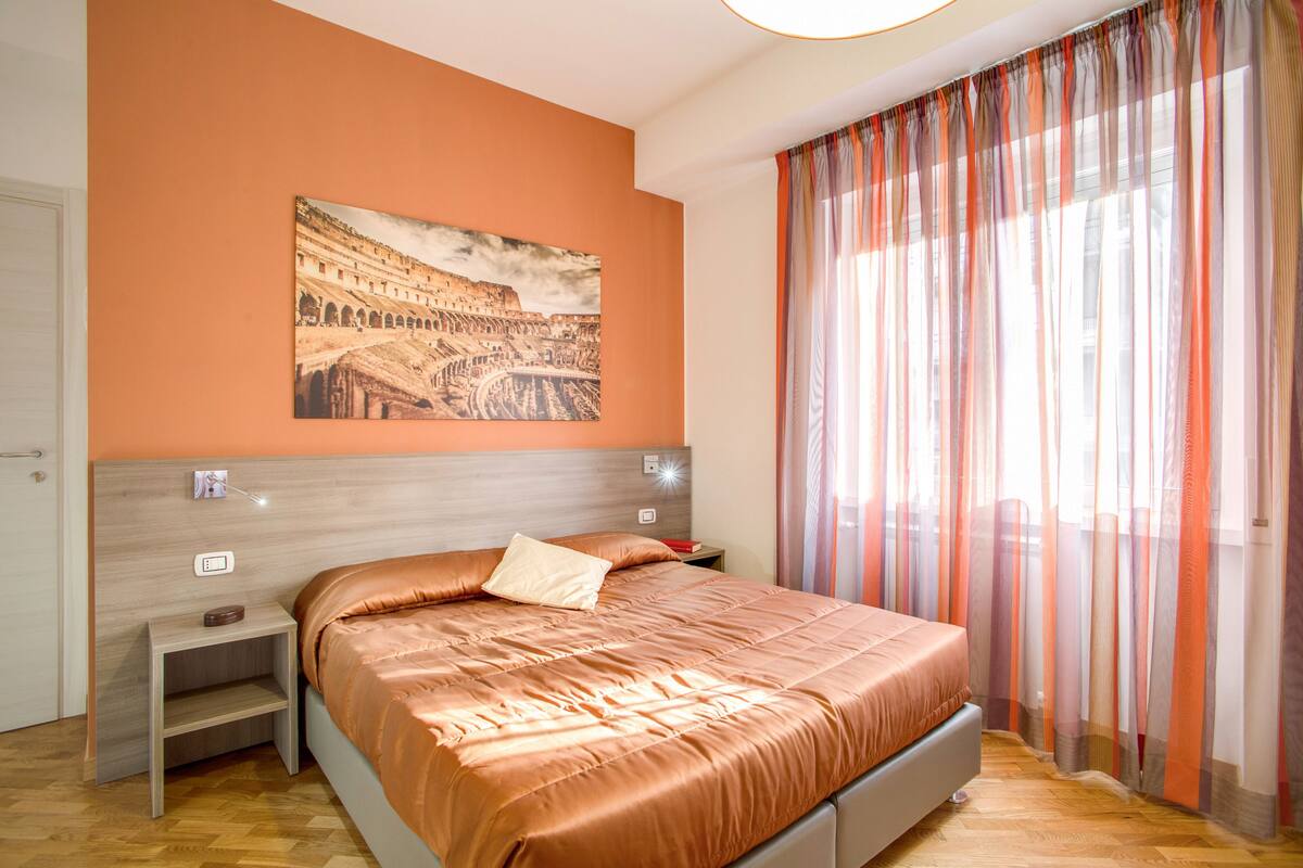 A bedroom with a bed, a nightstand, a wall-mounted lamp, and a window with sheer curtains.