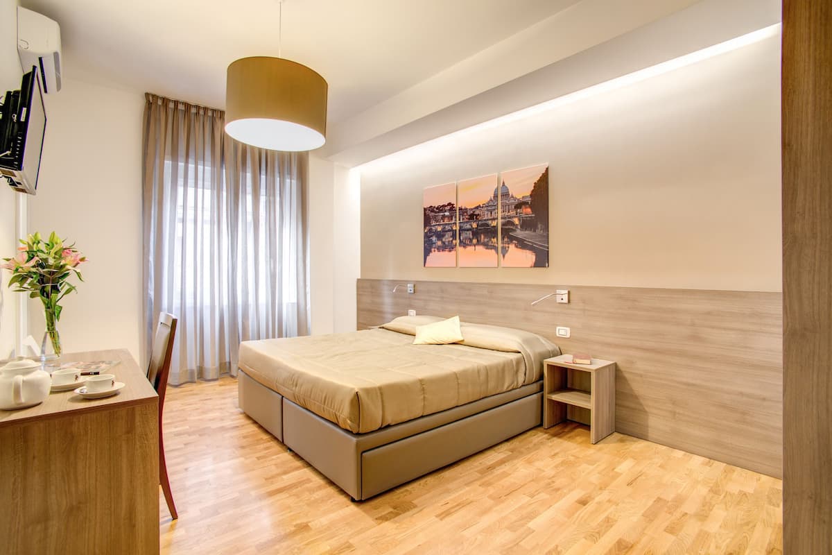A modern hotel room with a large bed, bedside tables, a desk with a chair, a vase with flowers, and a painting on the wall.