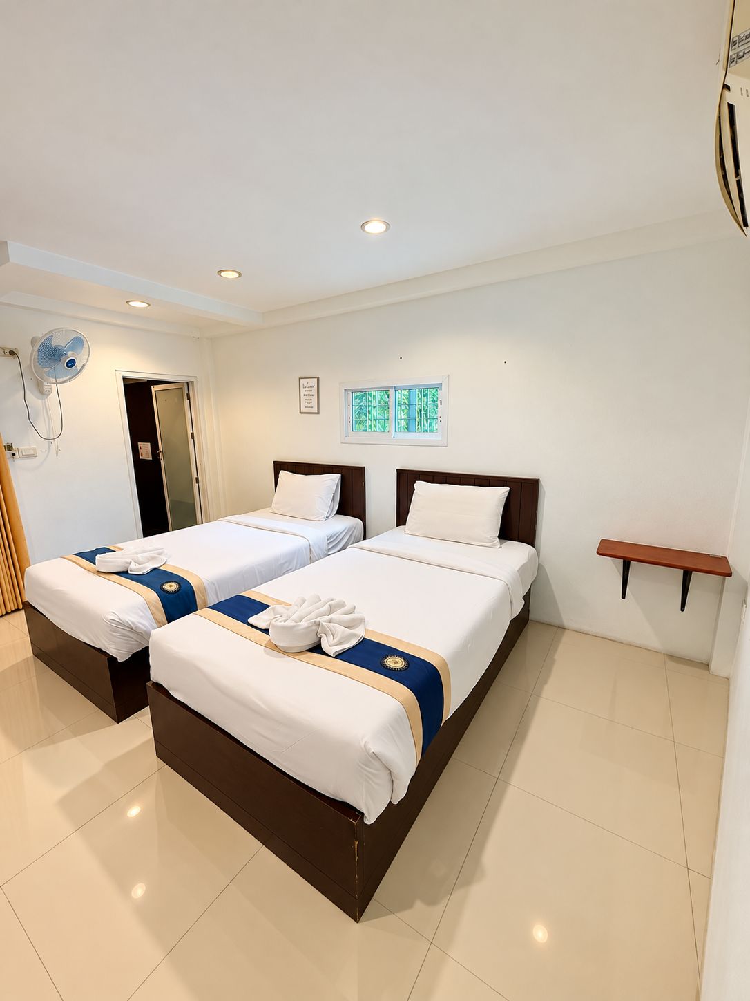 Ein Hotelzimmer mit zwei Betten, einem Deckenventilator, einem kleinen Fenster und einem an der Wand befestigten Regal.
