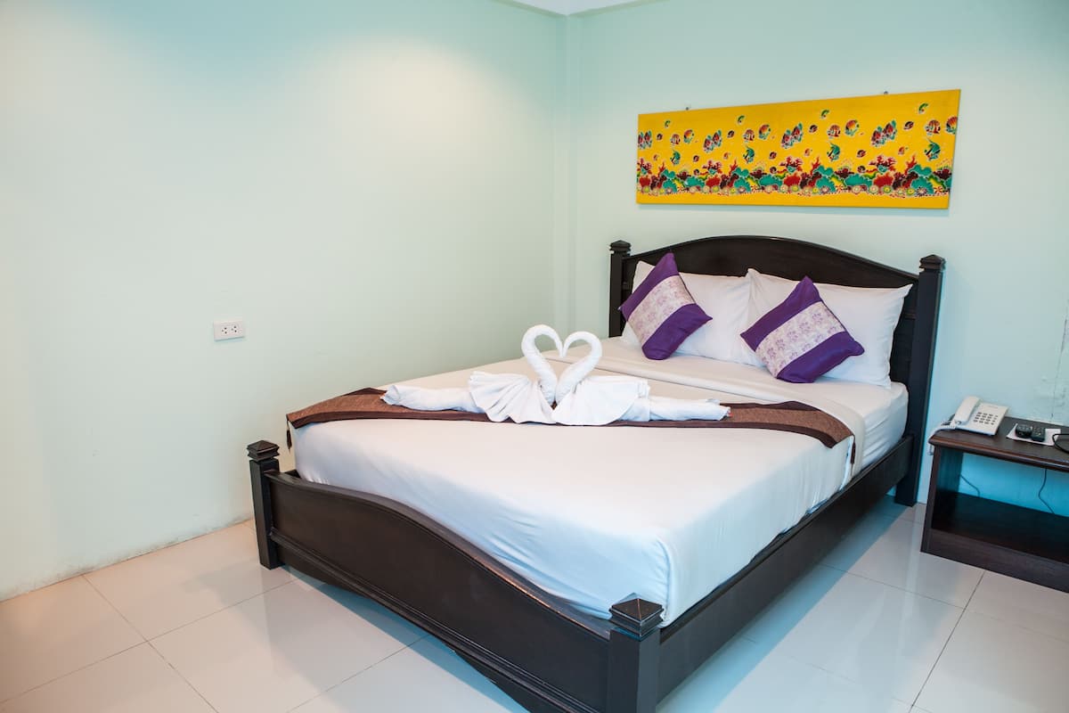 Una cama bien hecha con sábanas blancas y almohadas decorativas, una mesita de noche con un teléfono, y un cuadro abstracto de colores en la pared.