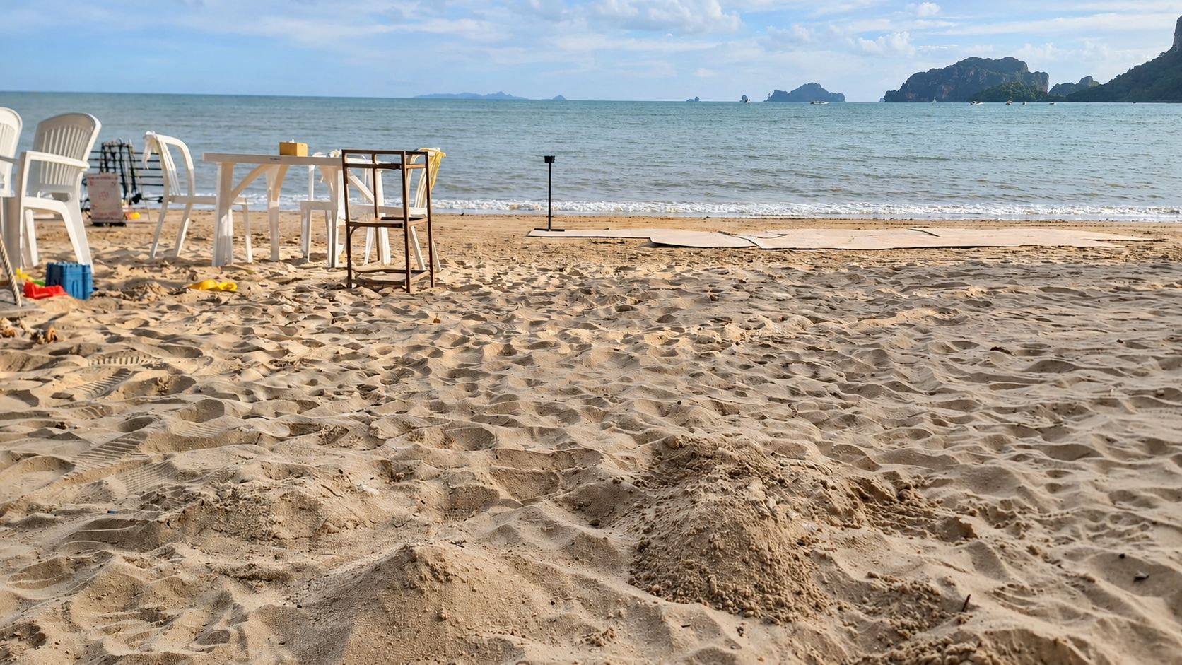 Una playa con sillas y mesas de plástico blanco, una pequeña mesa con un objeto amarillo y un poste negro.