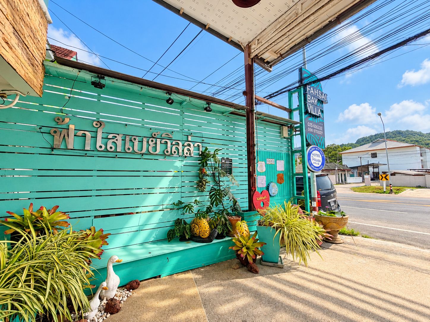 A hotel entrance with a green facade, a signboard with Thai script, and a variety of potted plants.