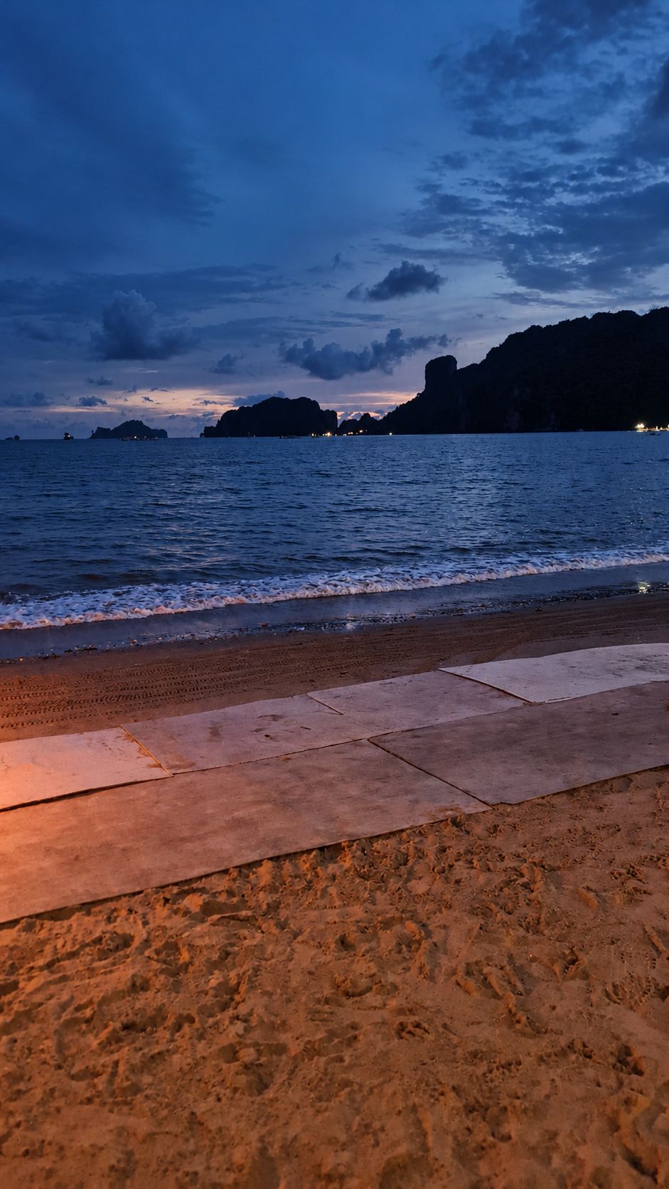 A beach with a paved walkway leading to the water, calm sea, and distant rocky formations under a twilight sky.