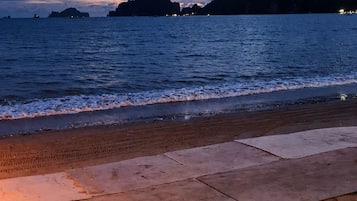 Una playa con un paseo marítimo empedrado que lleva al agua, mar tranquilo y formaciones rocosas distantes bajo un cielo crepuscular.
