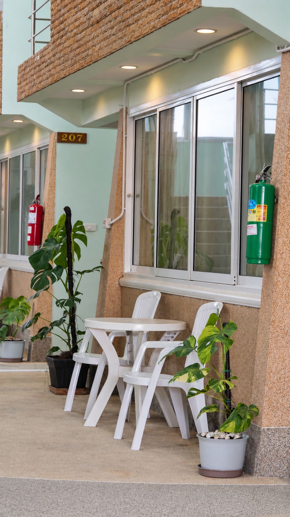 Una habitación de hotel con balcón, ventana con cortinas, extintor y plantas en macetas.