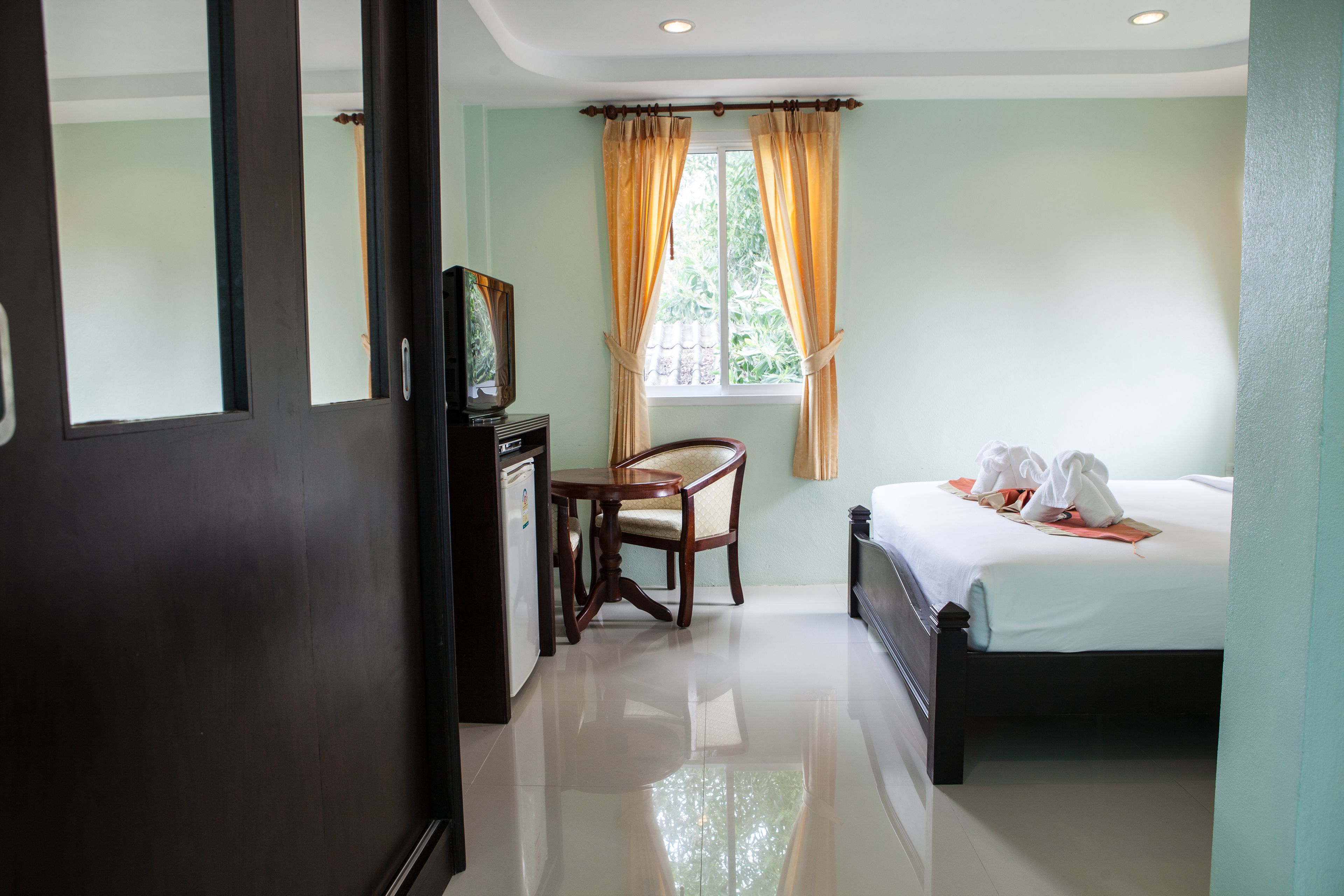 A hotel room with a bed, a desk with a chair, a window with curtains, and a refrigerator.