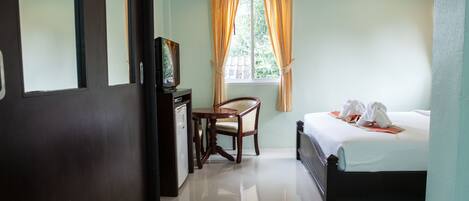 A hotel room with a bed, a desk with a chair, a window with curtains, and a refrigerator.