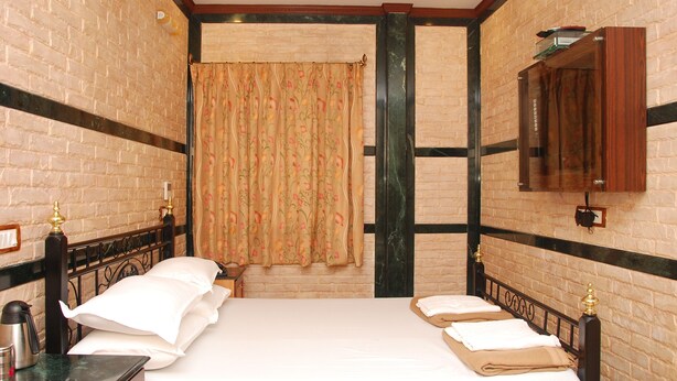 Hotelkamer met een bakstenen muur, een groot bed met witte lakens, een houten nachtkastje, een spiegel en een raam met bloemen gordijnen.