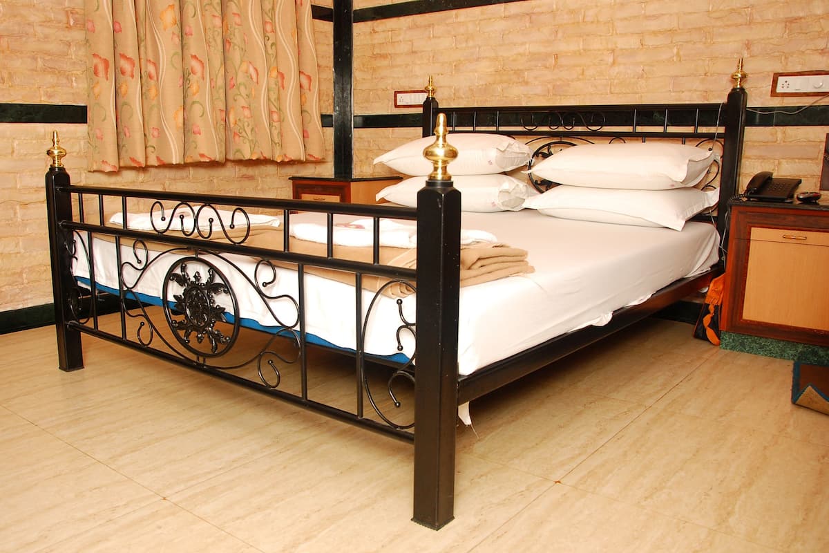 A bed with a metal frame, white bedding, and a wooden headboard. There is a nightstand with a telephone and a lamp. The room has a brick wall and a window with curtains.