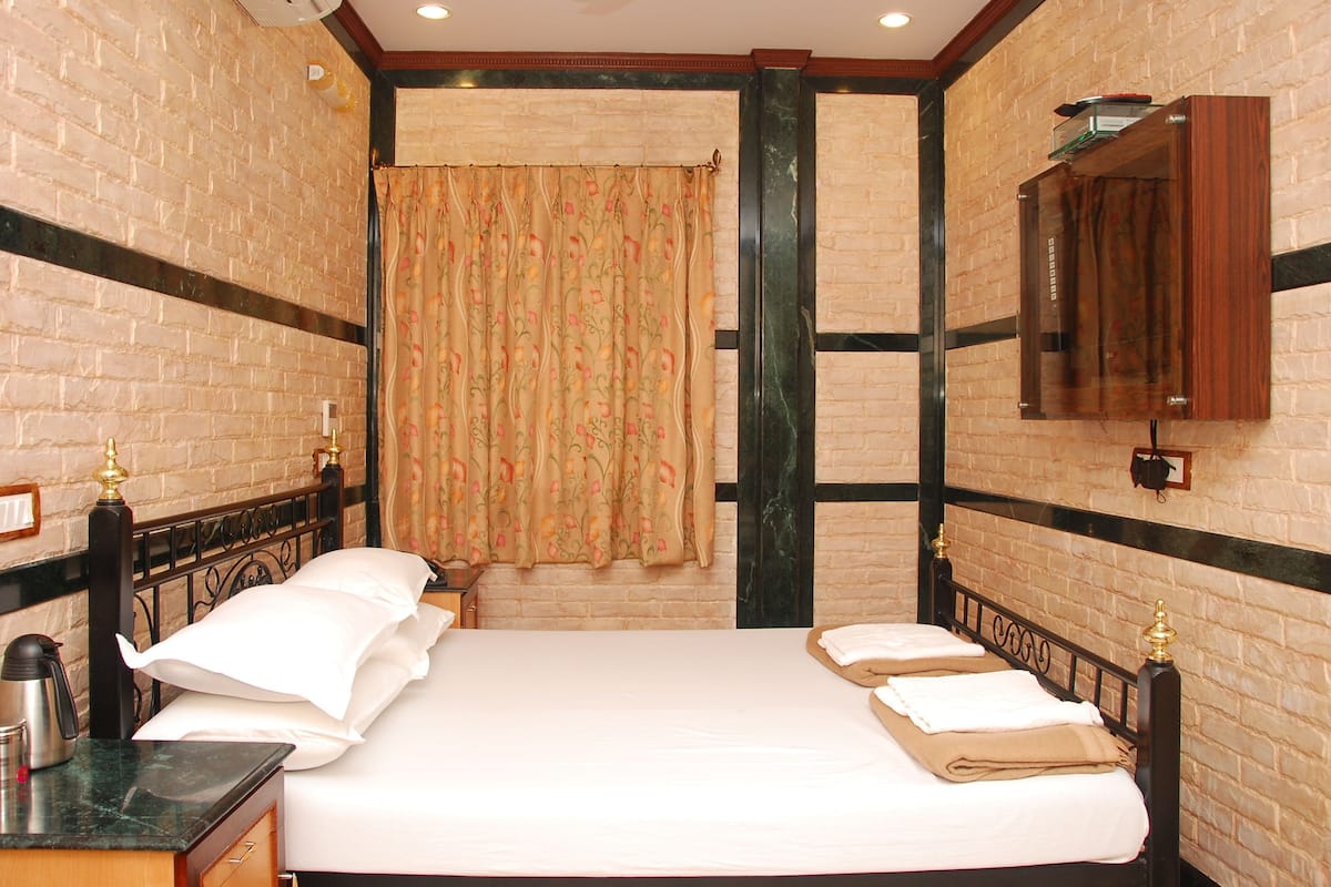 A hotel room with a brick wall, a large bed, a bedside table with a kettle, and a mirror.