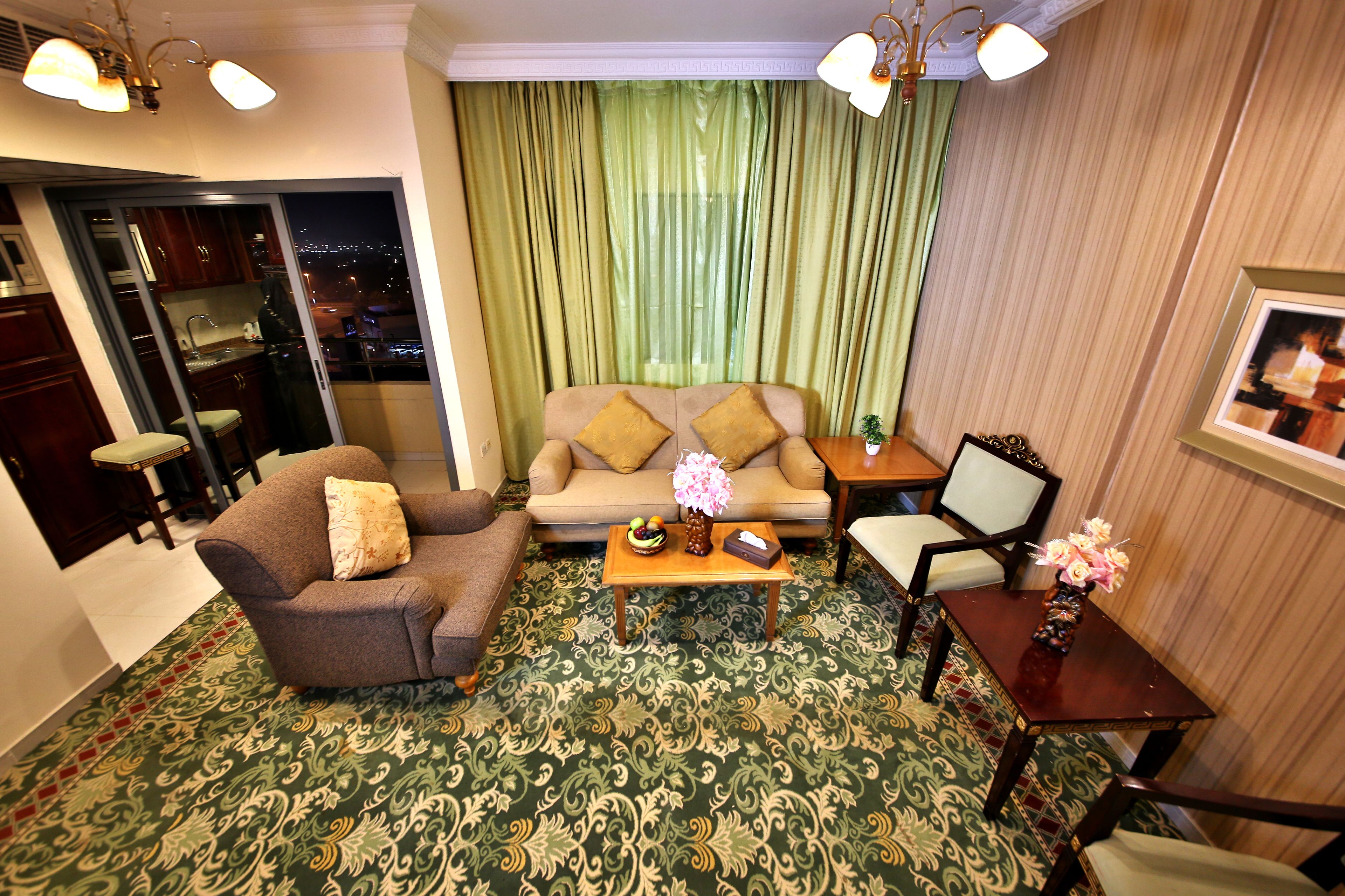 A living room with a sofa, armchairs, a coffee table, and a dining table with chairs. There is a view of the city at night through a window with green curtains.