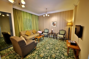 A hotel room with a green patterned carpet, a sofa, a chair, a small table with a vase, and a television mounted on the wall. - Midtown Furnished Apartments (Ajman)