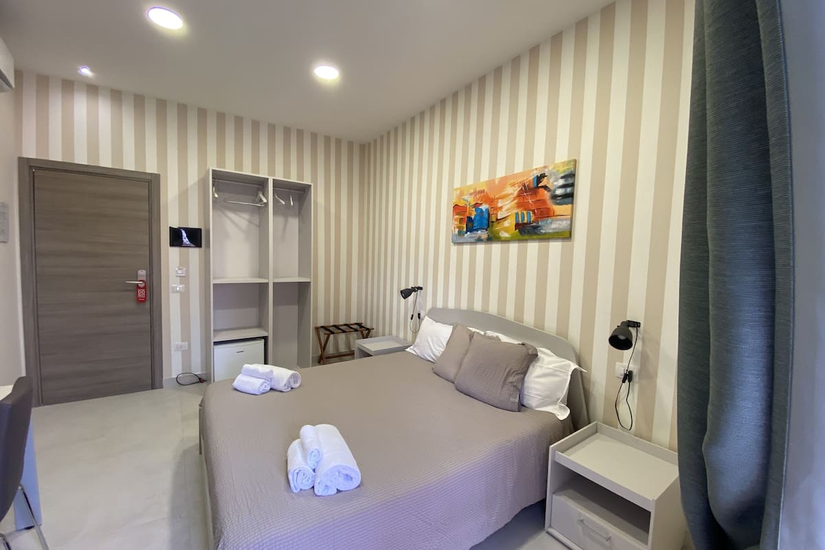 A hotel room with a bed, a wardrobe, a desk, and a painting on the wall.