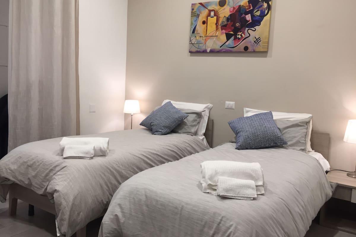 A hotel room with two beds, each with a pillow and a rolled towel. There is a painting on the wall above the beds.