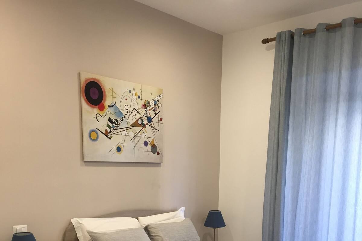 A hotel room with two beds, a painting on the wall, a lamp, and a window with curtains.