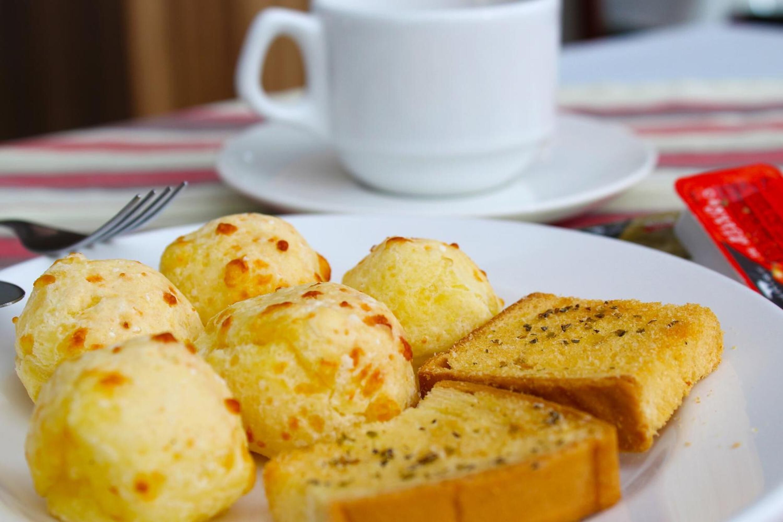 Un plato de comida con panecillos y una rebanada de pan, una taza blanca y un objeto rojo sobre una mesa.