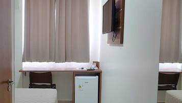 Una habitación de hotel con cama, escritorio con silla, televisor montado en la pared y un refrigerador pequeño.