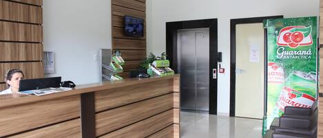 A reception desk with a wooden finish, a television mounted on the wall, and a refrigerator with a Guaraná Antarctica advertisement.