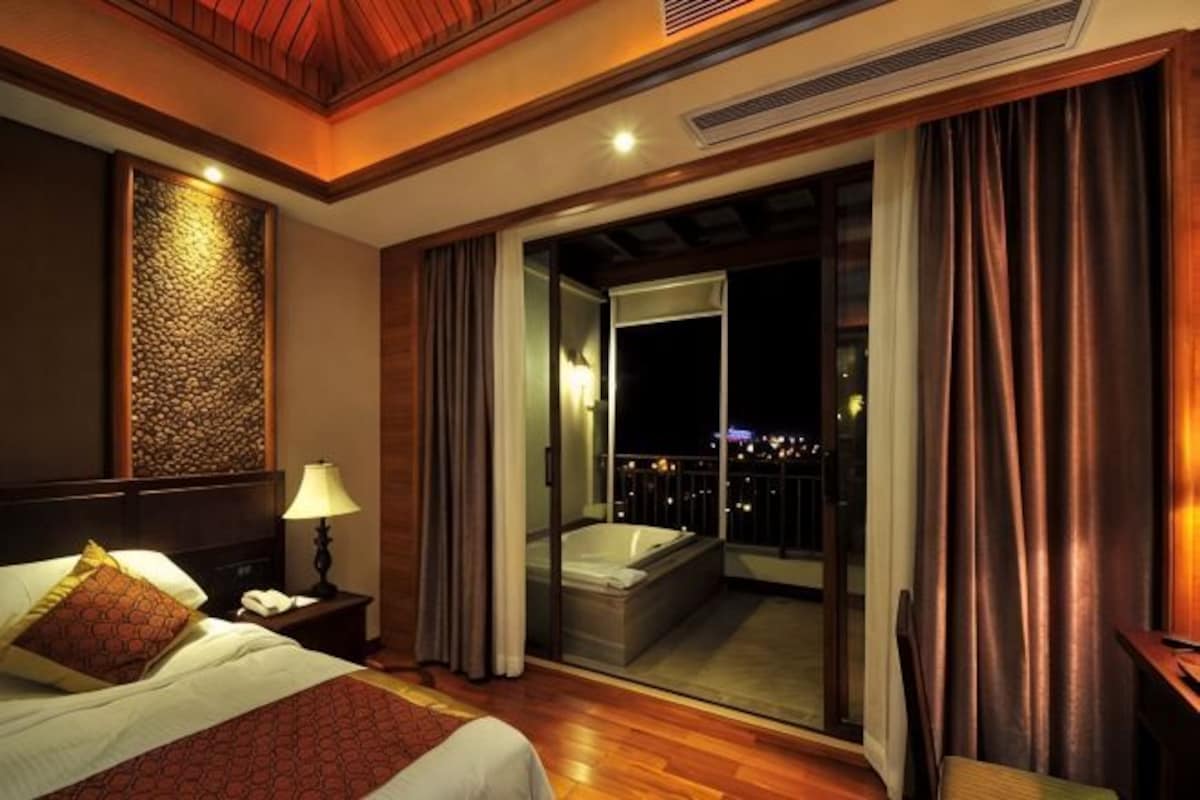 Habitación de hotel con cama, mesita de noche, baño con bañera y balcón con cortinas.