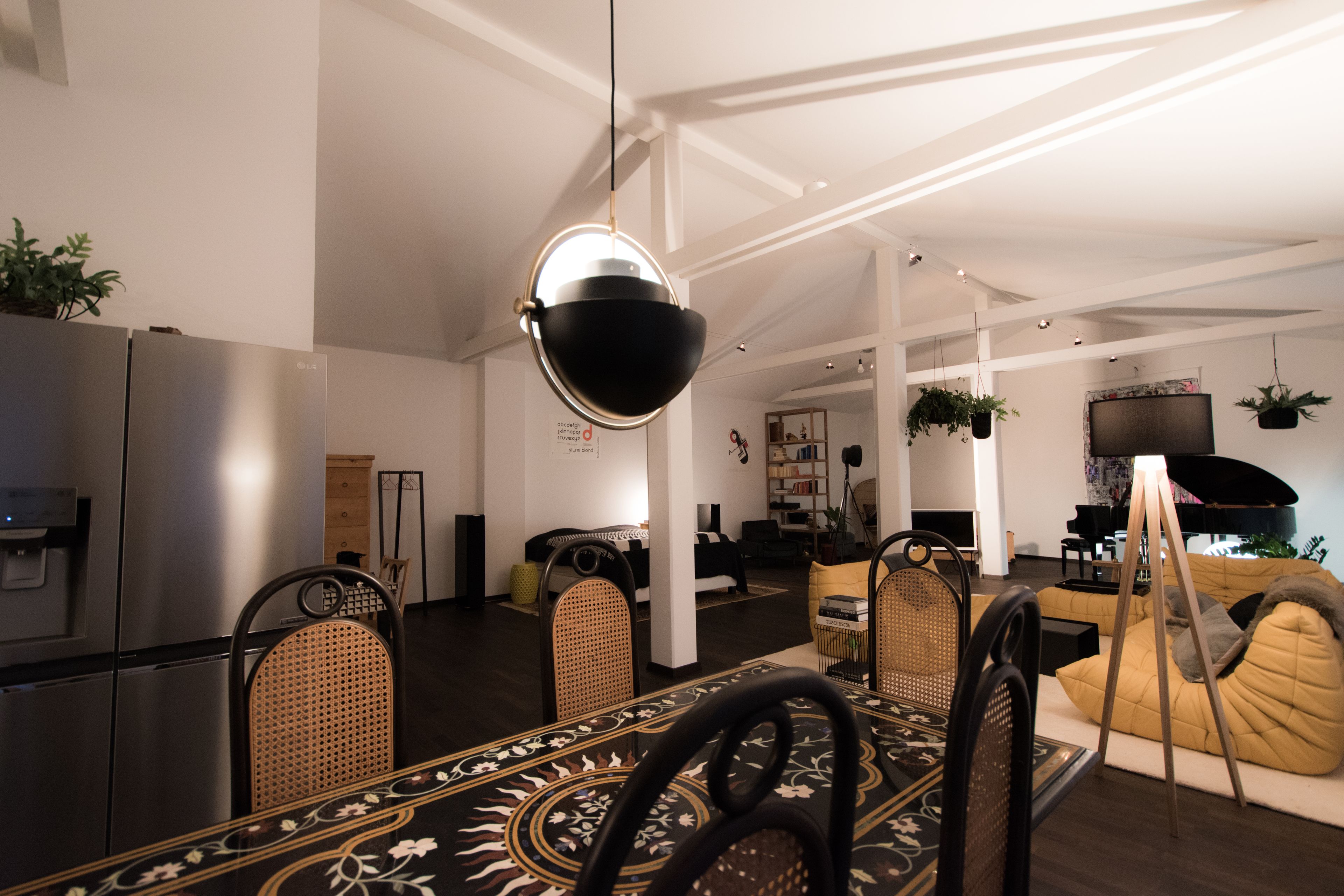 A modern living room with a dining area, featuring a large black bowl hanging light fixture, a piano, and a sofa.